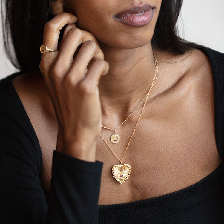A woman in a black off-shoulder top showcases two gold necklaces—one with a small round pendant, another with a large heart-shaped pendant—paired with the Solid Gold Quhier Que Demain Signet Ring.