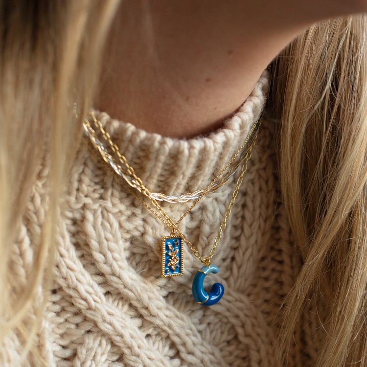 Close-up of a person in a chunky cream knit sweater wearing two gold necklaces—one is the Bubble Letter Necklace, 14k gold plated with a blue rectangular pendant; the other has a blue and gold swirl pendant. Long blonde hair is partially visible.