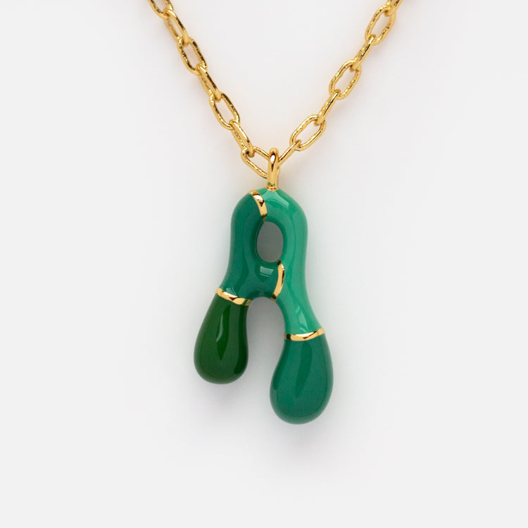 The Bubble Letter Necklace is a 14k gold plated piece with a green, abstract-shaped pendant featuring two rounded drops and slim gold accents, shown against a white background.