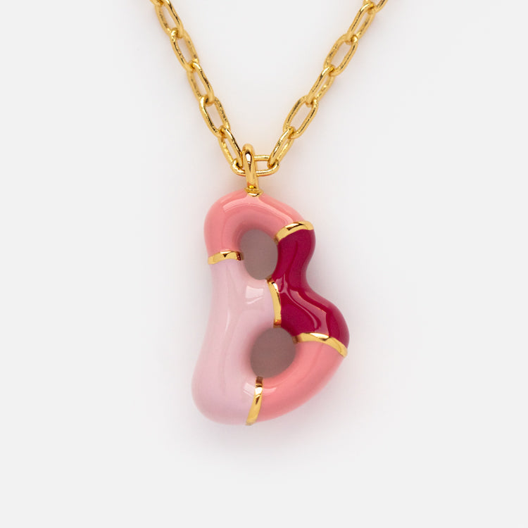 The Bubble Letter Necklace is a 14k gold plated piece with intertwined pink and red abstract shapes, gold accents, and small circular cutouts on the pendant, set against a white background.