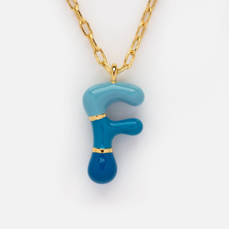 The Bubble Letter Necklace features a 14k gold plated chain with a glossy, smooth blue and teal glass pendant shaped like the letter F, accented in gold for a playful yet elegant look.