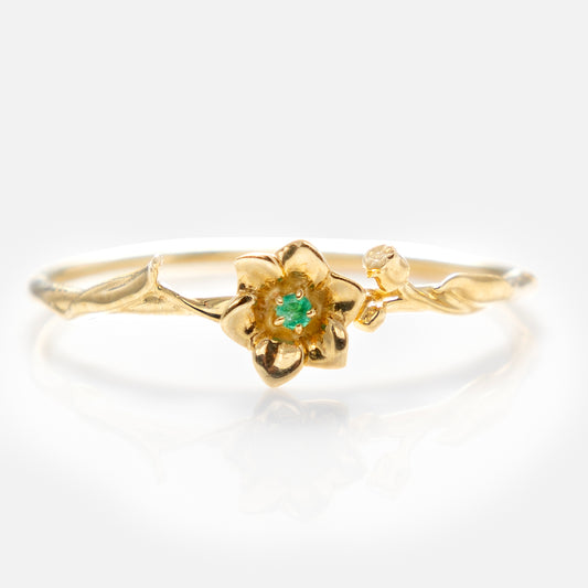 The Solid Gold Birth Flower Ring features a delicate flower design in solid gold, with a small green gemstone at the center and a tiny accent stone on the band, set against a white background.