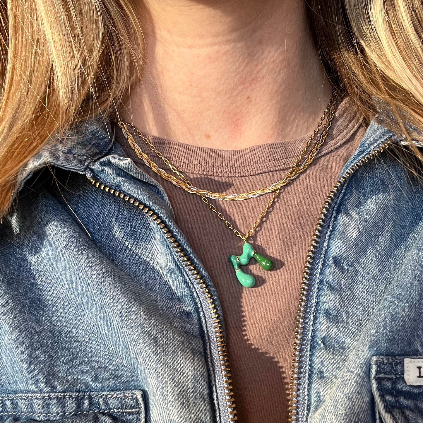 A person wearing a denim jacket over a mauve shirt, styled with two layered gold necklaces—one being the Bubble Letter Necklace—captured in natural sunlight.