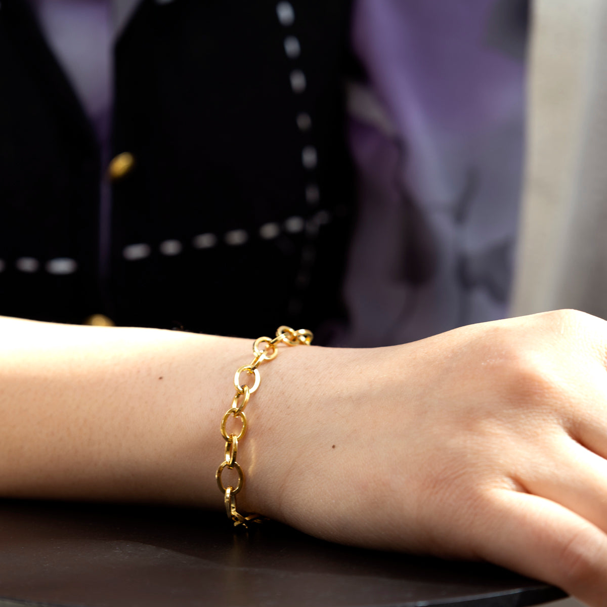A person wearing the Becca Bracelet, a gold chain link piece, rests their arm on a dark surface. They are dressed in a black and purple top with white stitching.