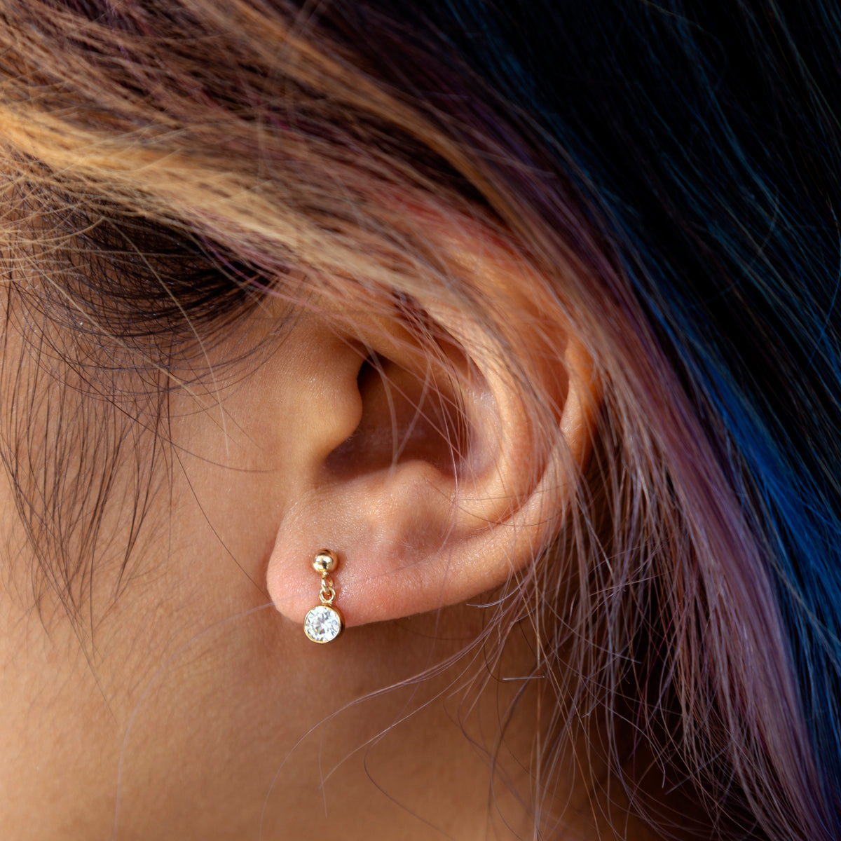 Close-up of a person’s ear with colorful, streaked hair, wearing the Billie Earrings Sample—delicate 14k yellow gold filled studs with a single round, dangling CZ gemstone.