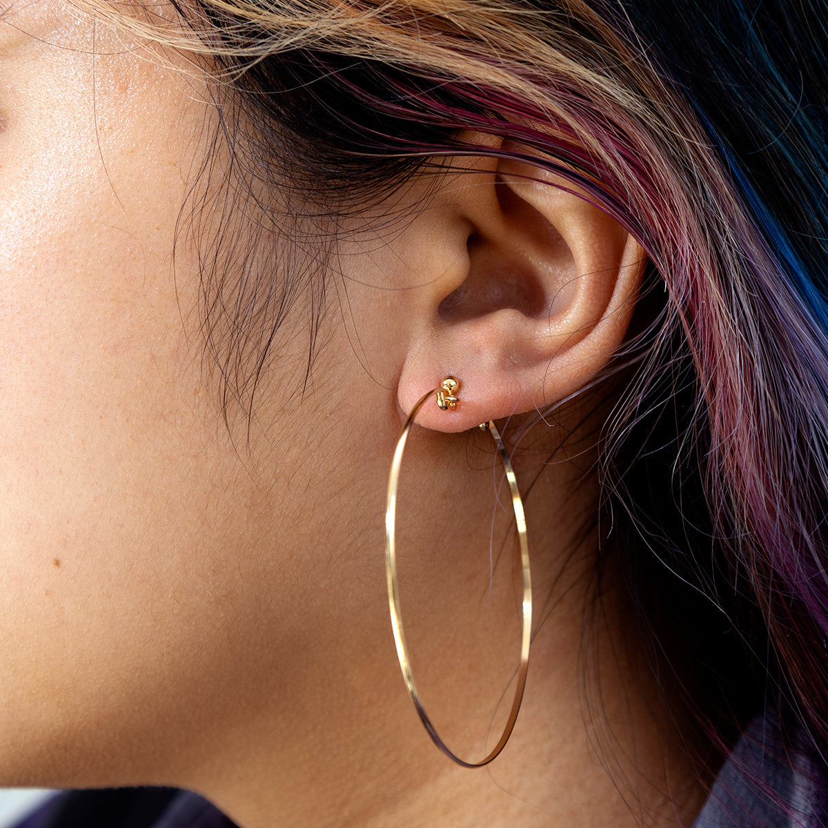 Close-up of a person's ear wearing Valour Hoops Sample, a large gold hoop earring paired with a small 14k yellow gold stud; hair features black, brown, and streaks of purple and blue.