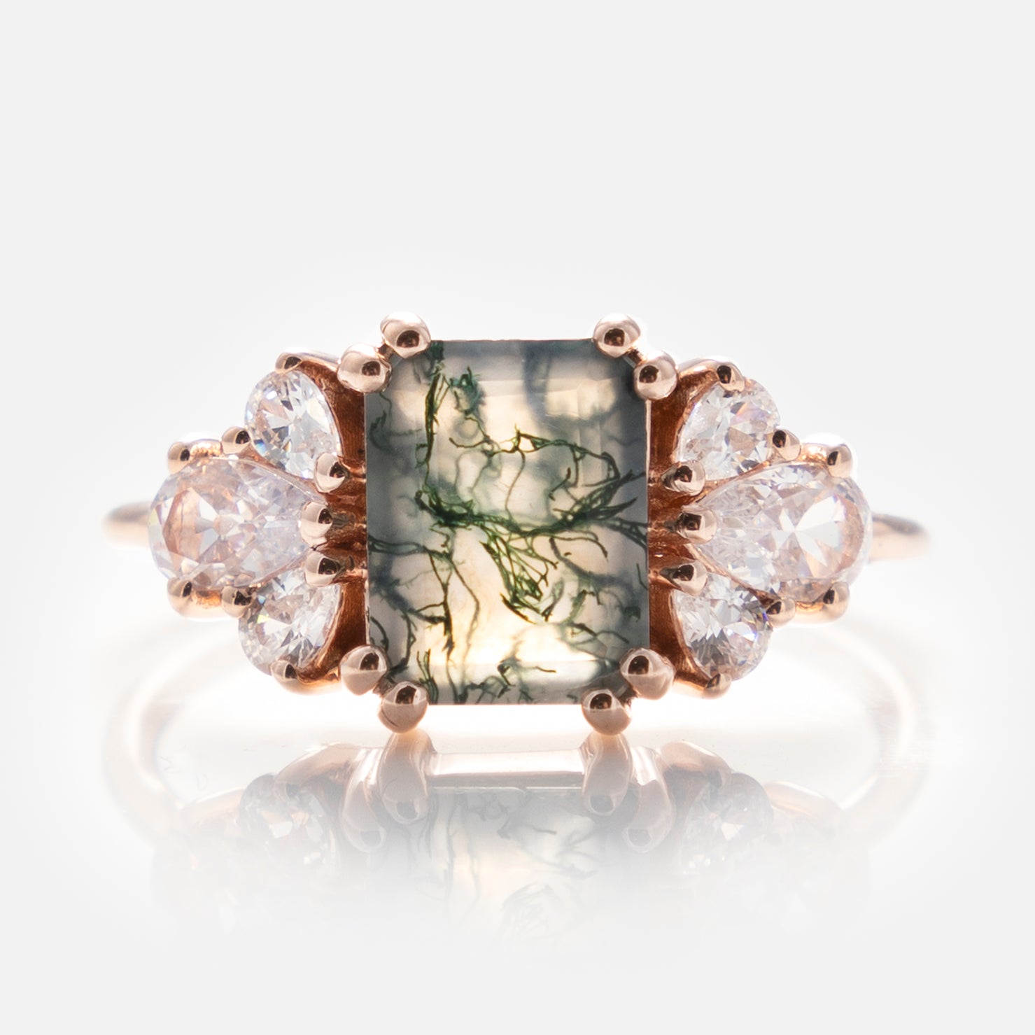 The Gemstone Empress Ring is a gold statement piece with a rectangular moss agate center stone featuring green inclusions, accented by clusters of three clear pear-shaped stones on each side, shown on a white background.