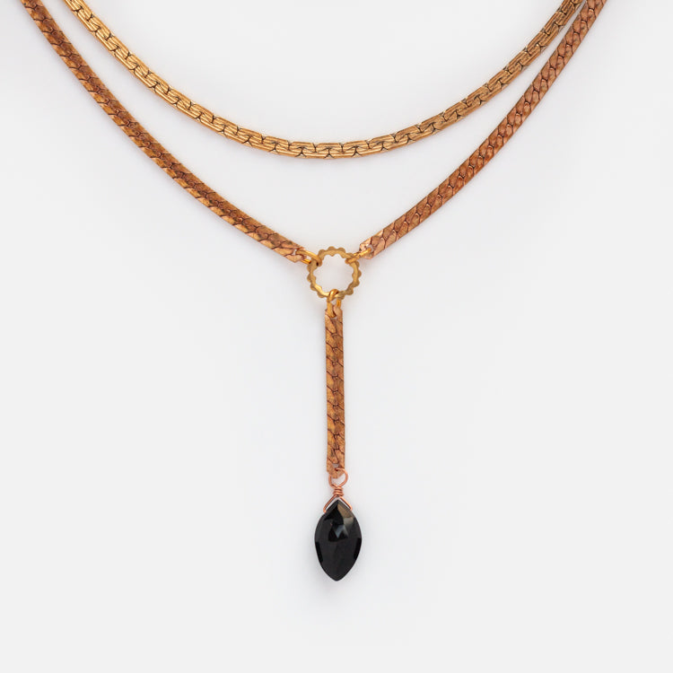 The Palmer Gemstone Necklace features a gold-layered recycled brass chain with a circular centerpiece and a single black teardrop pendant, creating an elegant, symmetrical design against a white background.