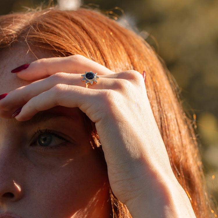 A close-up of a red-haired person holding their hand to their forehead, highlighting the Solid Gold Starburst Ring for Inspiration with a dark stone. The softly blurred outdoor background adds warmth and focuses on the jewelry.