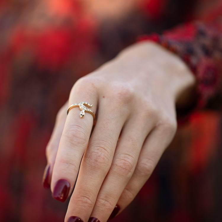 A close-up of a hand with dark red nail polish wearing the Solid Gold Diamond Snake Ring for Protection, set against a blurred background with rich red and dark tones.