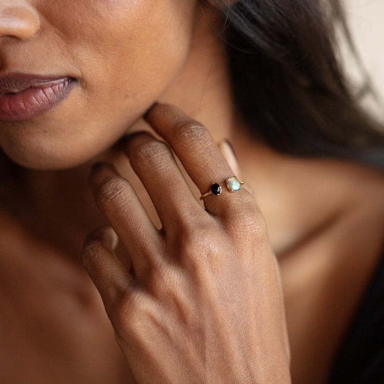 A close-up of a woman with brown skin and dark hair wearing a black top showcases the Solid Gold Open Front Ring for Protection, featuring a black and an iridescent labradorite stone set in 9k yellow gold.