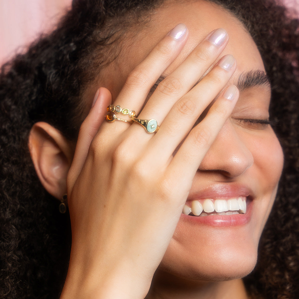 A smiling person with curly hair covers one eye with their hand, flaunting the Solid Gold Crystal Signet Ring for Manifestation. Neatly manicured nails and gemstone accents showcase their love for manifestation jewelry.