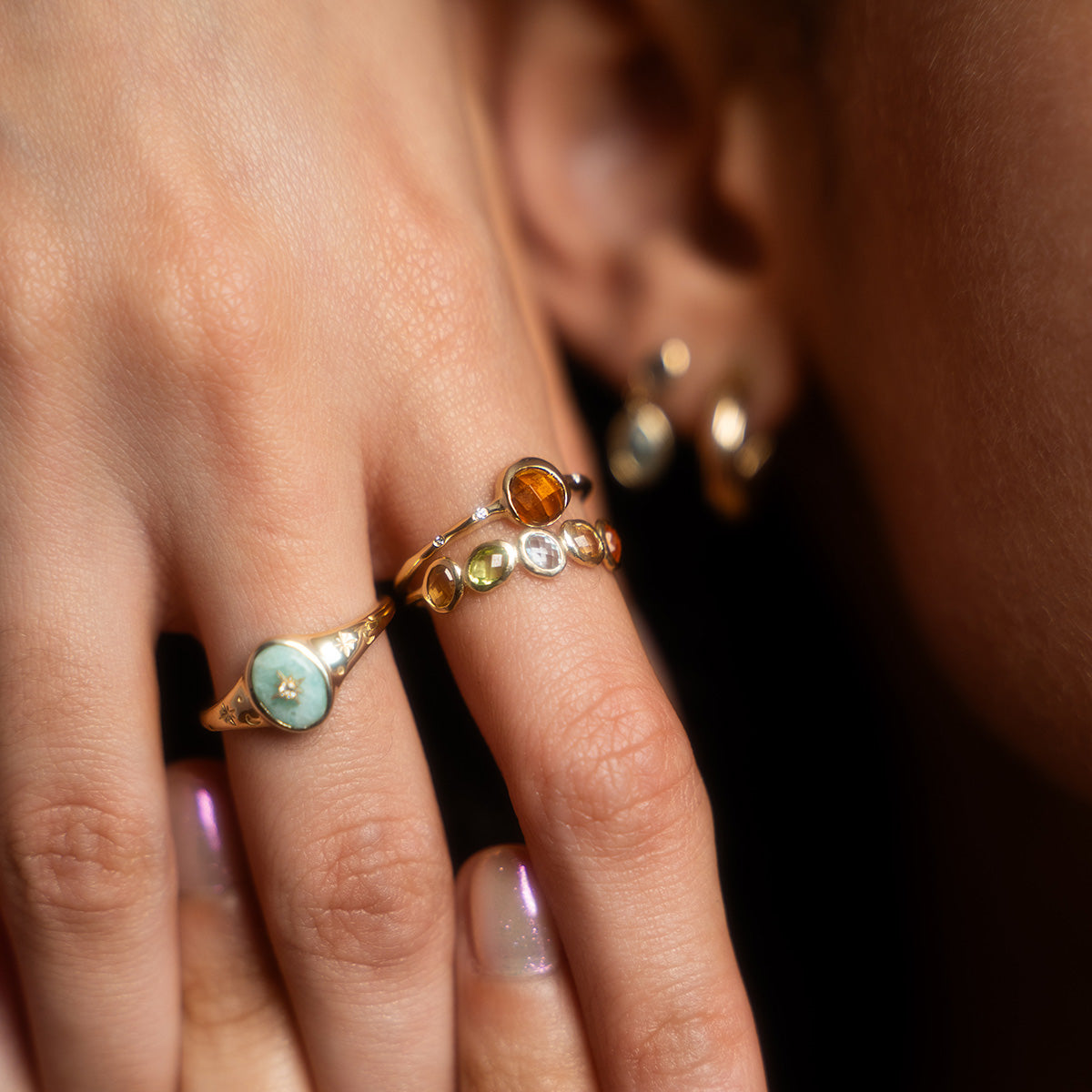 A close-up of a hand with warm skin tone and light pink nails wearing the Solid Gold Crystal Signet Ring for Manifestation, alongside two other gold rings, held near an ear adorned with gold and gemstone earrings.