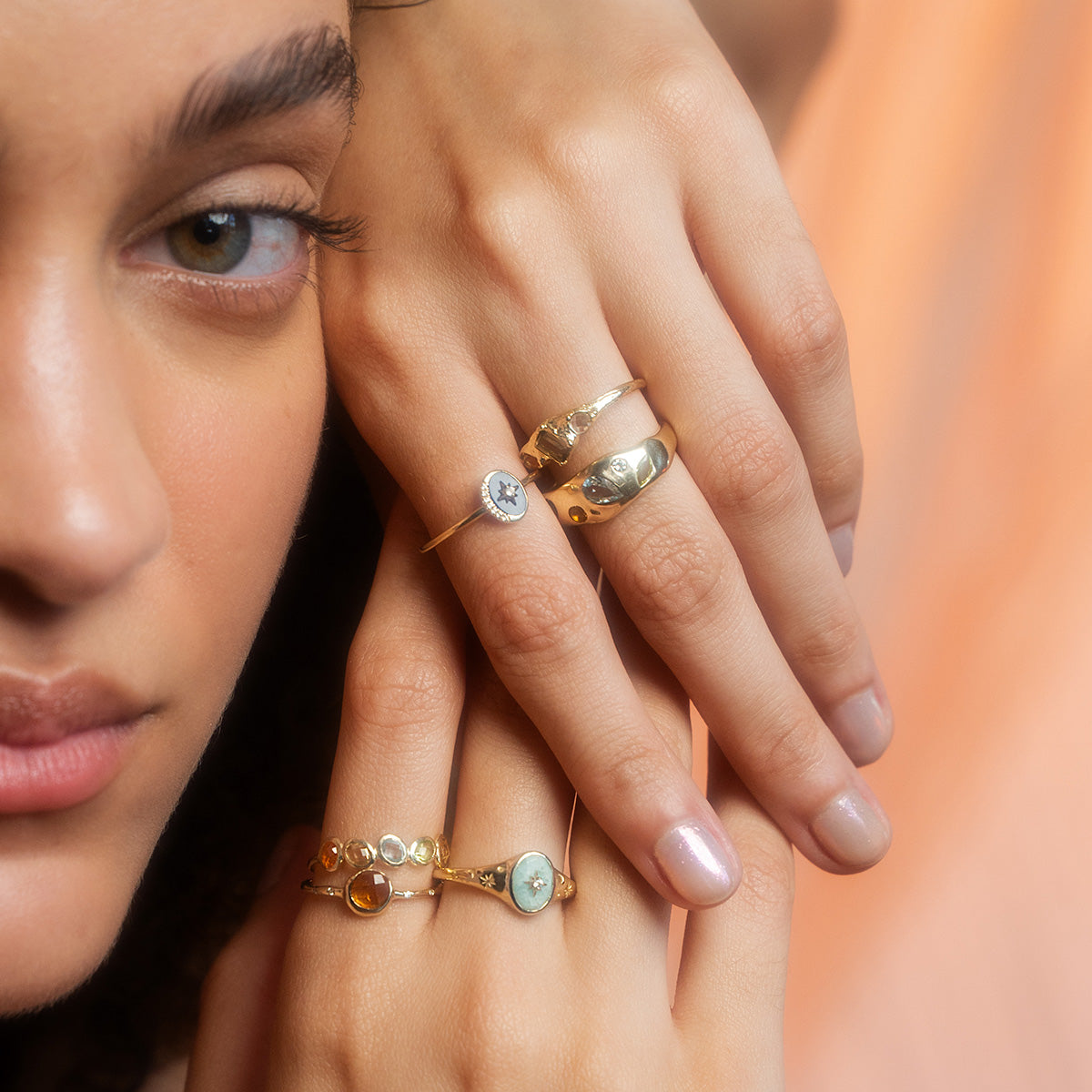 A close-up of a woman’s hands wearing the Solid Gold Crystal Signet Ring for Manifestation, accented with pearls and decorative gold, focuses on her eye and softly glowing skin.