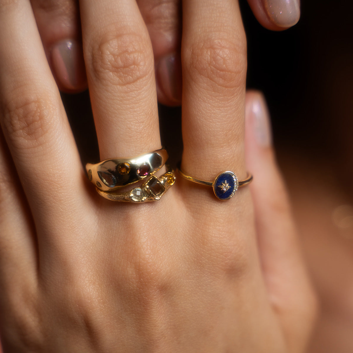 A close-up of a hand with three gold rings, including the Solid Gold Bold Organic Dawn Band, a wavy ring, and a Local Eclectic exclusive with a blue circular stone featuring a small star pattern. Fingernails are neatly manicured.