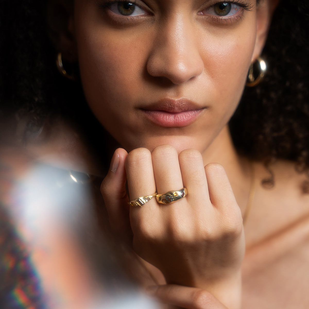 A woman with curly hair and gold hoop earrings poses in soft light, her hand near her face displaying two chunky rings—one being the Solid Gold Bold Organic Dawn Band. Her expression is calm and serene.