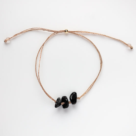 The Solid Gold Mantra Bracelet features three irregular black stones on a thin, light brown adjustable cord—a delicate natural crystal piece ideal for meditation jewelry. Shown on a white background.