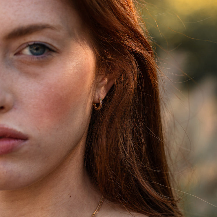 Close-up of a woman with fair skin and red hair, wearing Solid Gold Live by the Sun Rainbow Huggie Hoops, gazing forward with a serious expression. Warm sunlight softly blurs the background. Jewelry by Harvest Moon Local Eclectic.