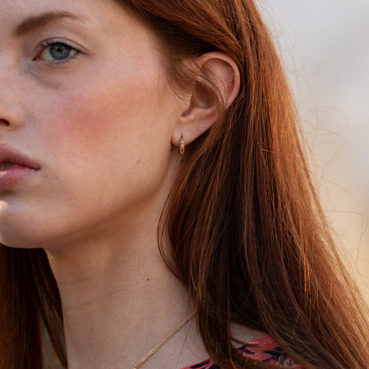 A close-up of a woman with long red hair, light skin, and blue eyes wearing Solid Gold Live by the Sun Rainbow Huggie Hoops and a gold necklace, looking slightly to the left.
