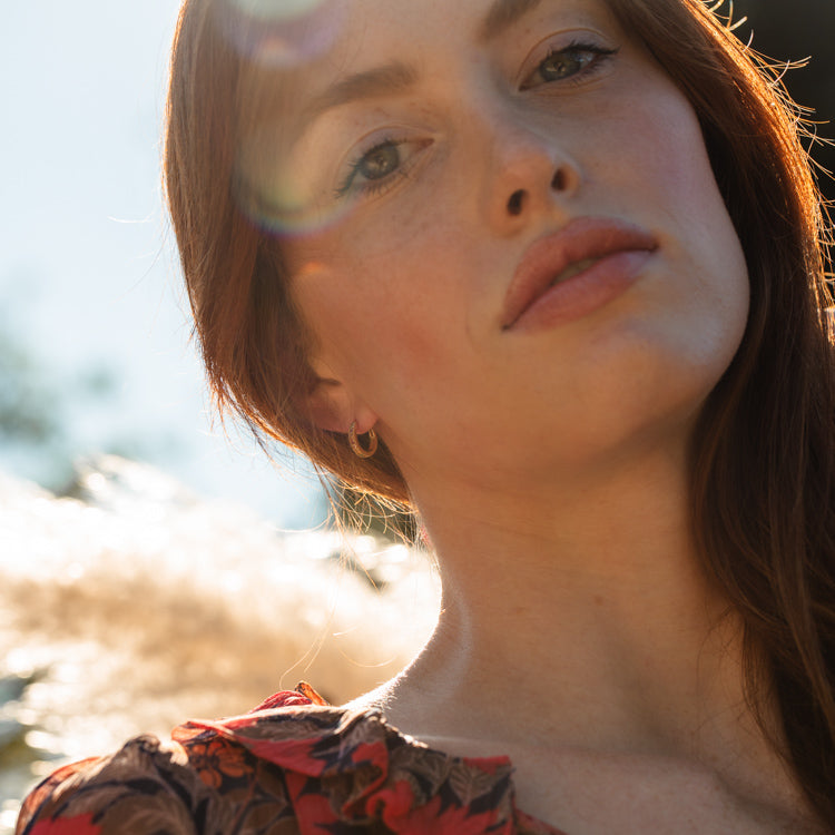 A woman with fair skin and long auburn hair gazes into the camera, wearing a floral top and Solid Gold Live by the Sun Rainbow Huggie Hoops from Harvest Moon Local Eclectic. Sunlight bathes her in a soft glow with lens flare in the background.