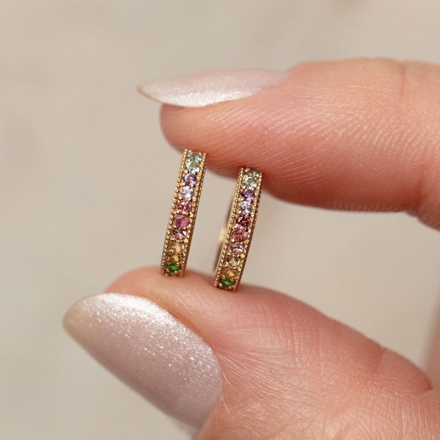 A hand with pale, shimmery nail polish holds two Solid Gold Live by the Sun Rainbow Huggie Hoops—gold earrings set with colorful gemstones in green, pink, purple, and blue.