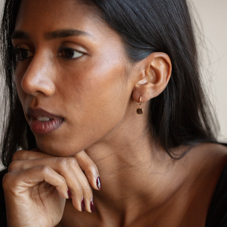 A woman with long dark hair and polished nails rests her chin on her hand, wearing Solid Gold Smoky Quartz Charm Huggies for Grounding. Her smooth brown skin and a neutral background highlight the earring’s elegance.