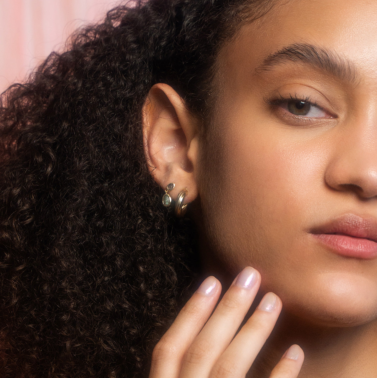 Close-up of a woman with curly hair wearing Solid Gold Labradorite and Quartz Drop Earrings for Protection, her pale pink manicured fingers gently resting on her cheek as she gazes calmly to the side.