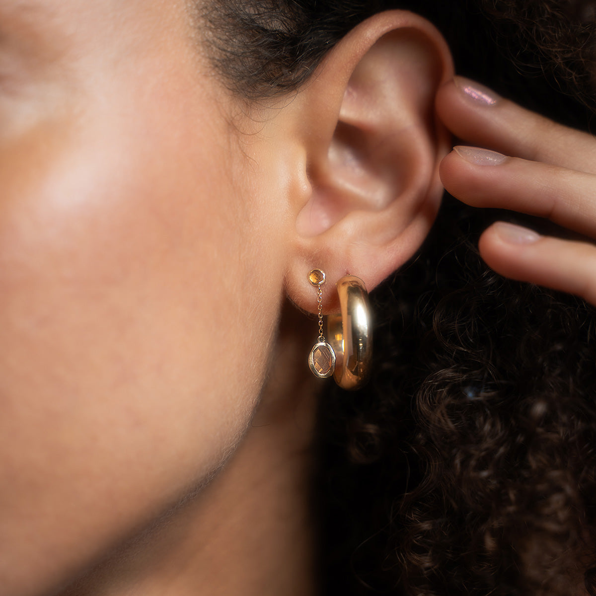 Close-up of a woman's ear wearing Solid Gold Orange Garnet and Sunstone Dusk Drop Earrings, featuring a small stud with a dangling gem and a large, thick gold hoop. Her hand, curly hair, and skin are partly visible.