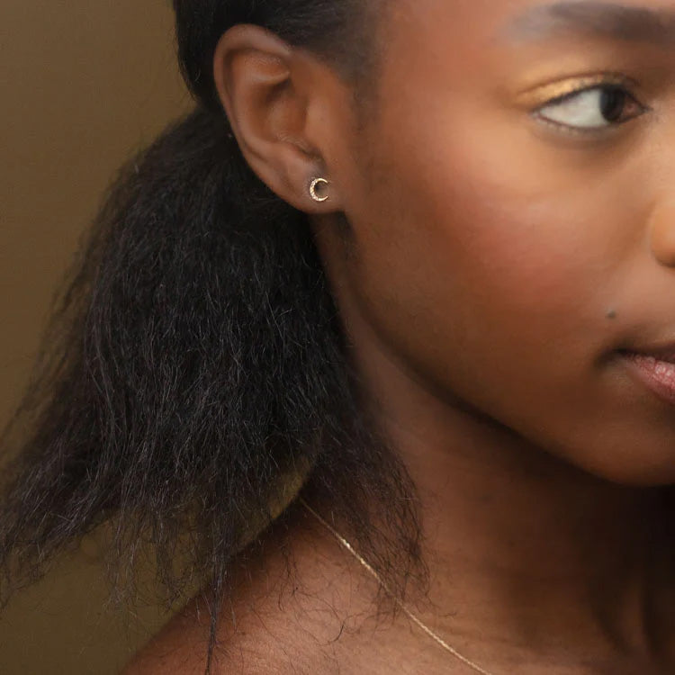 A close-up of a woman’s face and neck highlights her smooth skin, natural makeup, and dark hair, drawing attention to the Solid Gold Waxing and Waning Crescent Moon Studs—ideal for showcasing mindful jewelry.