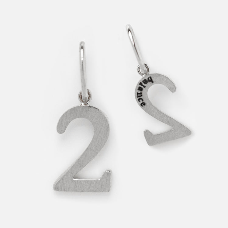 Two Solid Gold Number Charm earrings in White Gold, shaped as the number 2, one showing the front and the other the back with a subtle engraving on simple hooks. A unique personalized jewelry piece photographed on a plain light background.