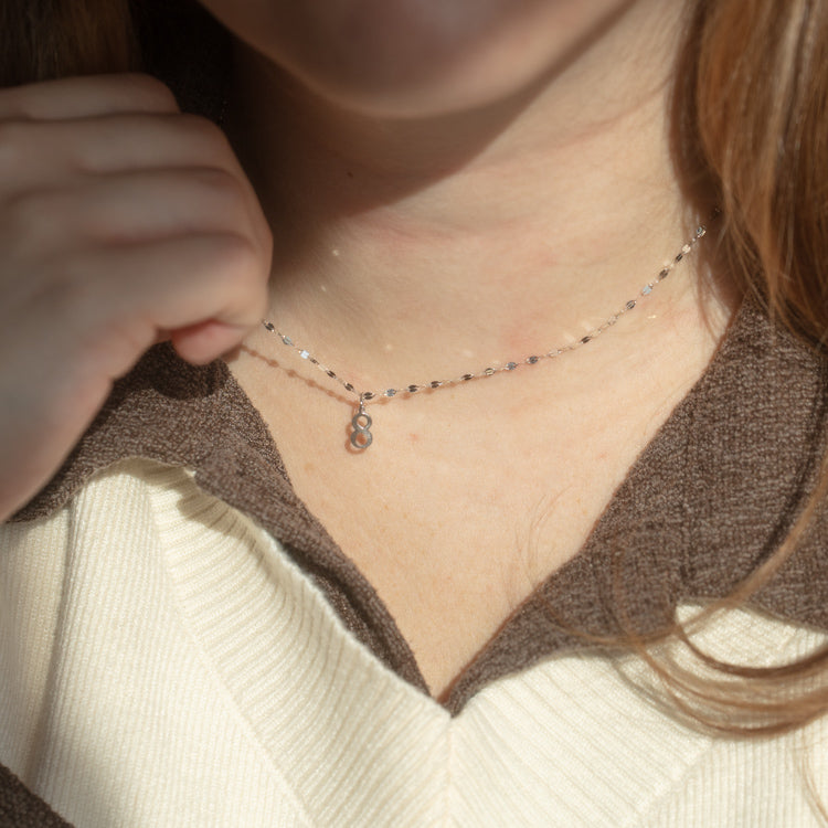 A person wearing a brown and cream sweater holds the Solid Gold Number Charm in White Gold, featuring a small number 8 pendant, shown close up against their neck and collarbone in natural light.