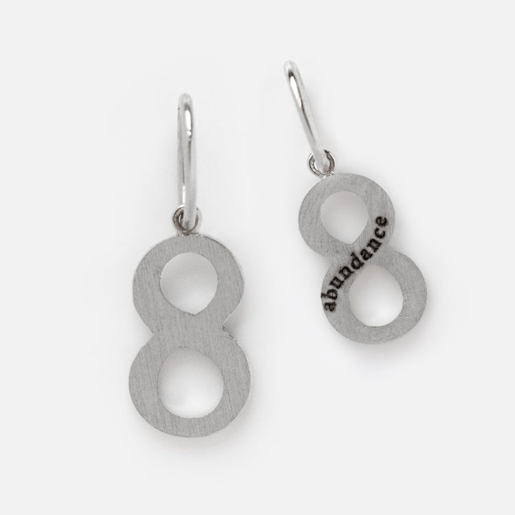The Solid Gold Number Charm in White Gold features earrings shaped like the number eight—one plain, one delicately engraved with abundance along the lower loop—for an elegant, personalized touch.