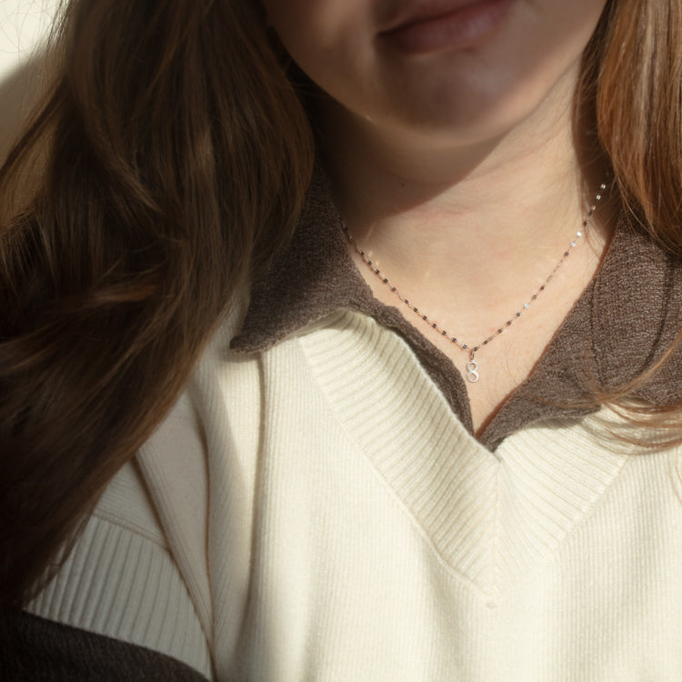 A woman with long brown hair wears a cream and brown collared sweater and a delicate Solid Gold Number Charm in White Gold necklace. Soft lighting highlights her neck and part of her face.
