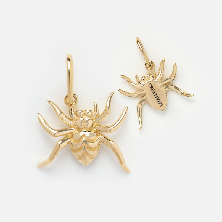 Two Solid Gold Nature Charms shaped like spiders are displayed—one shows its detailed front, the other is flipped to reveal CREATIVITY engraved on the back. Both charms feature attached loops for hanging.