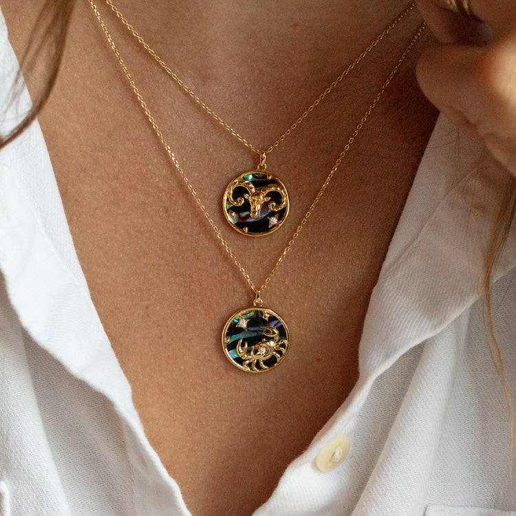 A person in a white shirt wears two layered necklaces: the Zodiac Pendant Necklace Aries Sample in 14k gold plate and a round pendant with intricate gold and dark designs, both elegantly resting on the chest.