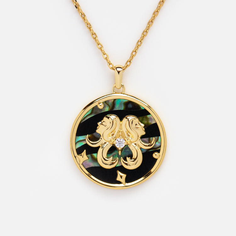 The Zodiac Pendant Necklace features a 14k gold plated round pendant with an ornate gold Gemini sign, a central diamond, and an abalone shell background with iridescent green and blue tones.