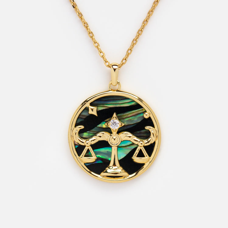The Zodiac Pendant Necklace features a 14k gold-plated round pendant with a scales of justice and star design, set against an iridescent abalone shell background.