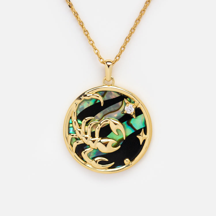 The Zodiac Pendant Necklace features a 14k gold plated round pendant with an intricate gold Scorpio symbol on iridescent abalone shell, accented by a sparkling gemstone for a celestial touch.