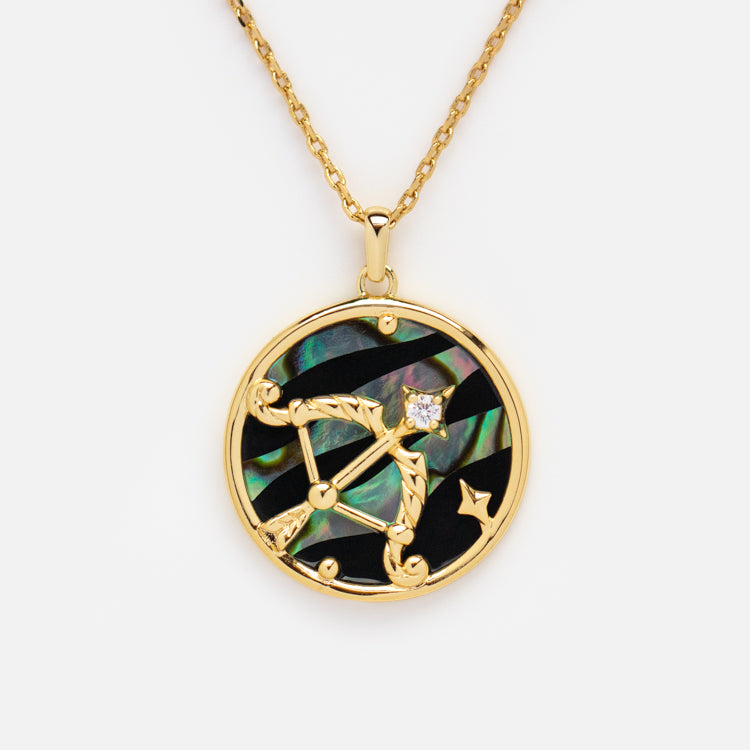 The Zodiac Pendant Necklace is a 14k gold plated piece with a round constellation pendant, bow and arrow design, iridescent abalone shell inlay, black accents, and a sparkling clear gemstone.
