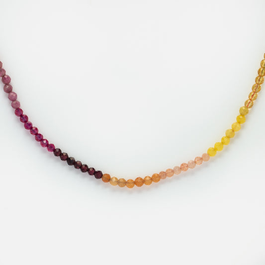 The Live by the Sun Rainbow Crystal Beaded Necklace is shown in a curved shape, with small round beads transitioning from deep purple and red to orange, pink, and yellow against a white background.