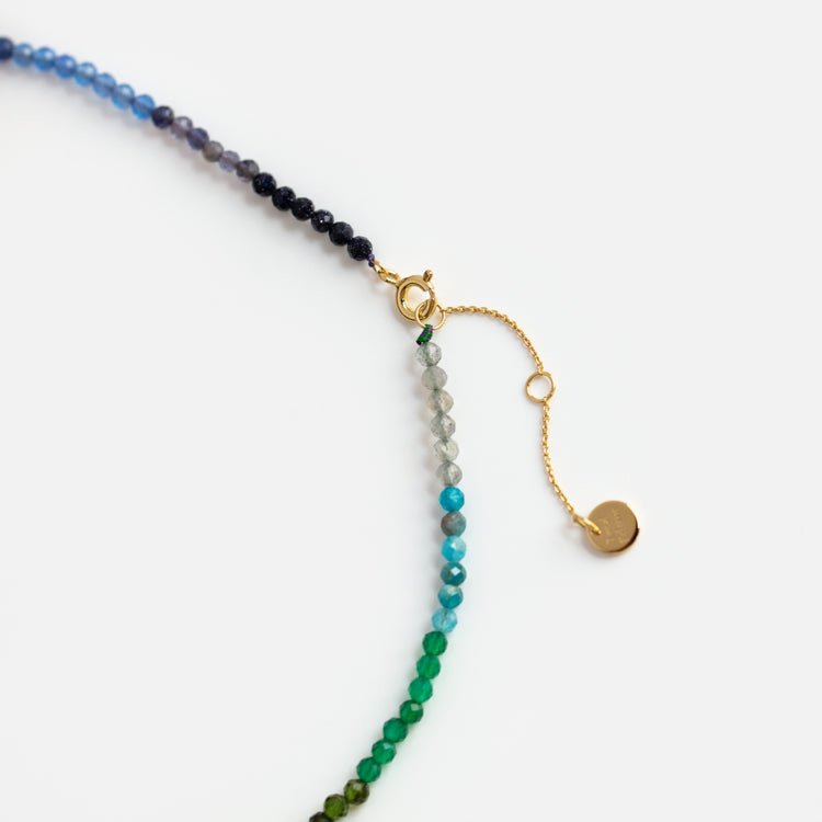 The Live by the Sun Rainbow Crystal Beaded Necklace features small, faceted natural crystals that fade from dark blue to green, 14k gold plating, an adjustable clasp and chain, and a round gold charm, displayed on a white background.