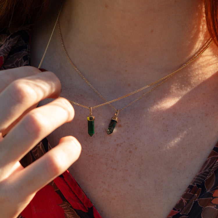 Close-up of a person with fair skin wearing two Solid Gold Crystal Charms for Manifestation, each featuring a green natural crystal pendant. They wear a red and black patterned top, with their hand partially visible near the necklaces.