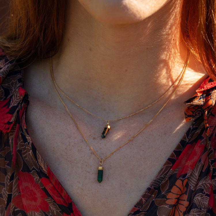 A person layers two Solid Gold Crystal Charm for Manifestation necklaces—each with green natural crystal pendants, one shorter than the other—over a red and brown floral blouse, ideal for those who believe in manifestation.