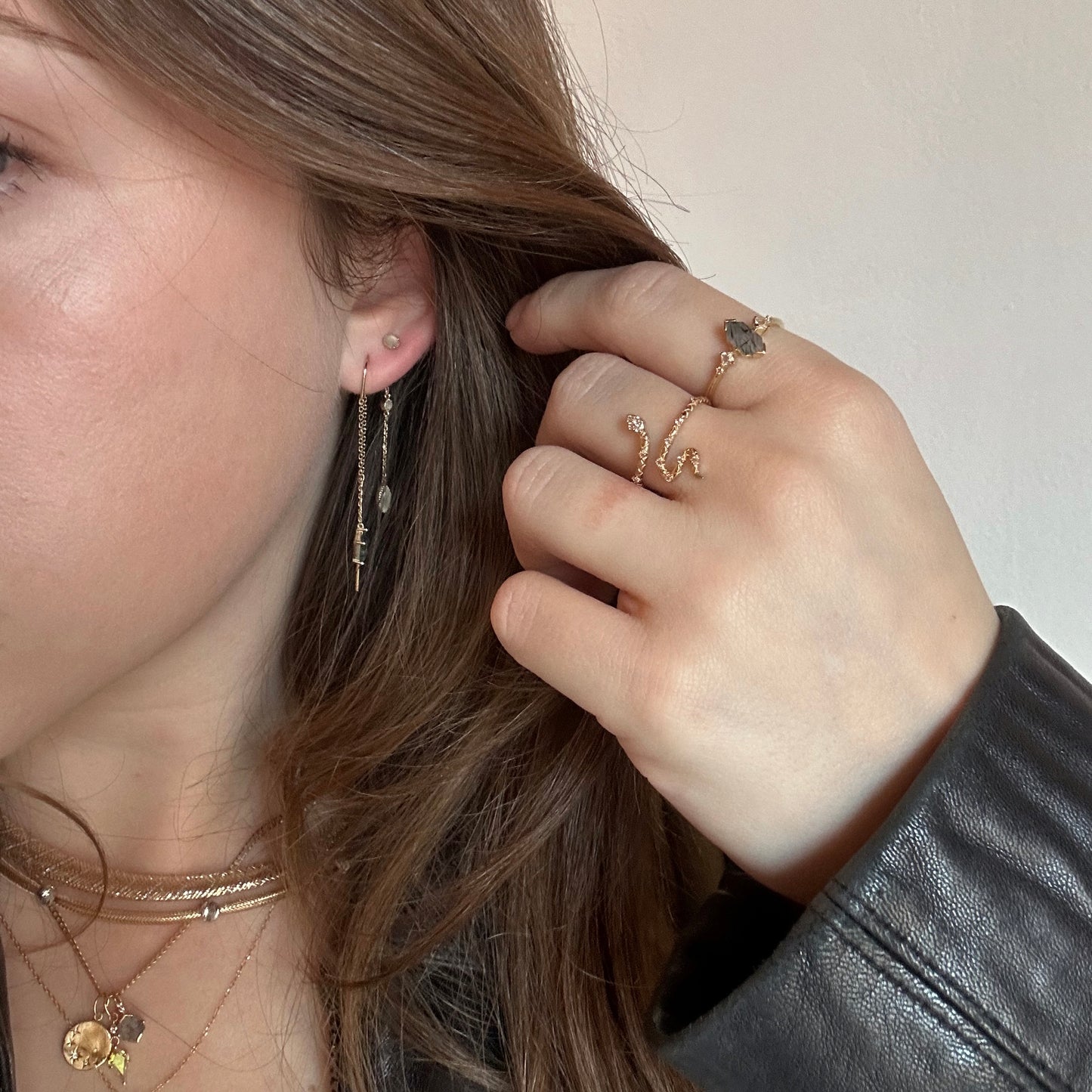 A woman wears layered necklaces, multiple rings, and Solid Gold Aquatic Agate Elements Kite Threader Earrings. She touches her hair with one hand while wearing a black leather jacket. The photo highlights her jewelry and accessories.