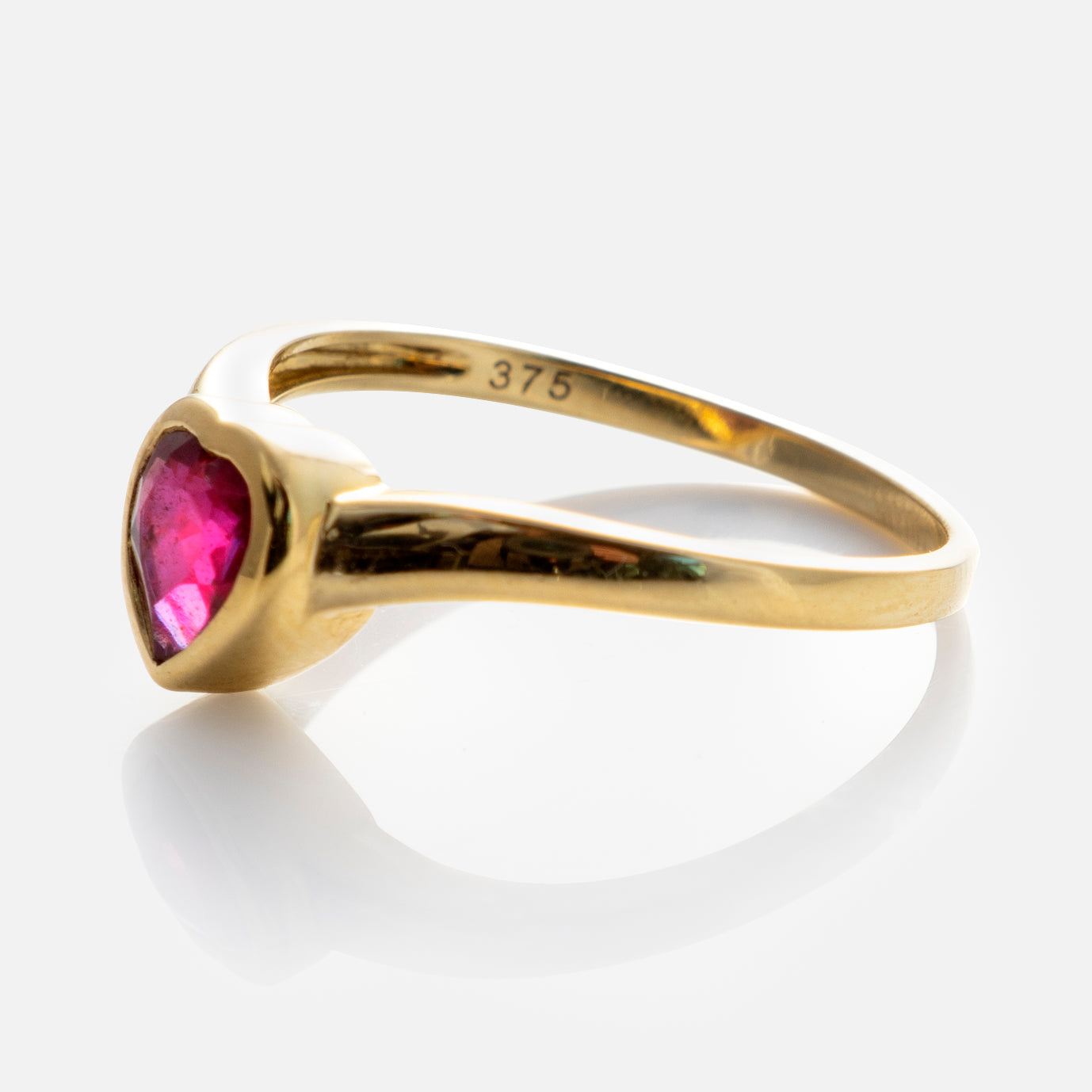 The Solid Gold Bezel Set Ruby Heart Ring, marked 375 inside the band, features a heart-shaped ruby and is displayed on a reflective white surface.
