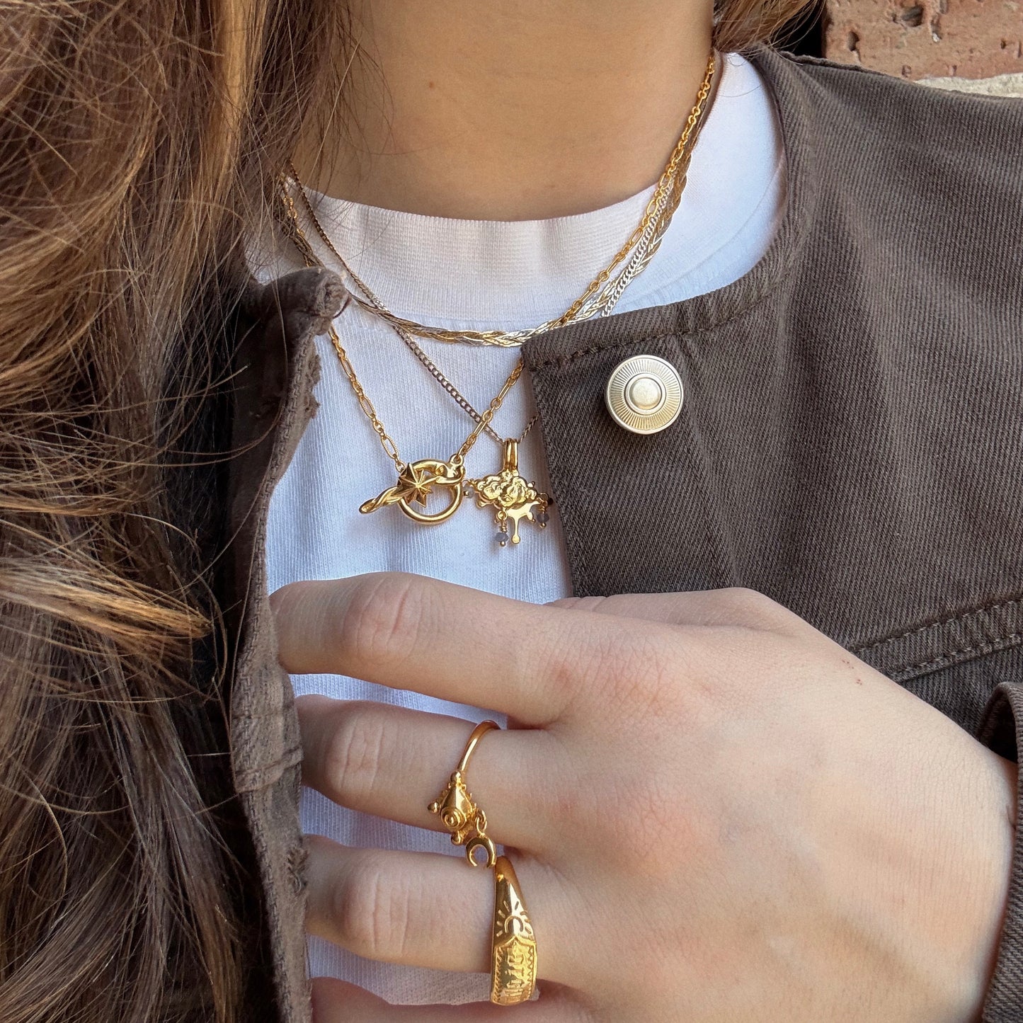 A person wears Charms by Hunt of Hounds layered gold necklaces and rings, styled with a white shirt, a brown jacket with a large button, and partially visible wavy brown hair.