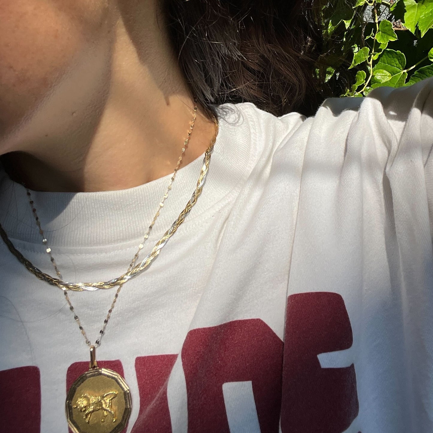A person wearing a white t-shirt with large red letters and a Solid Gold Mixed Metal Braided Necklace stands in sunlight, with green leaves and shadows in the background.
