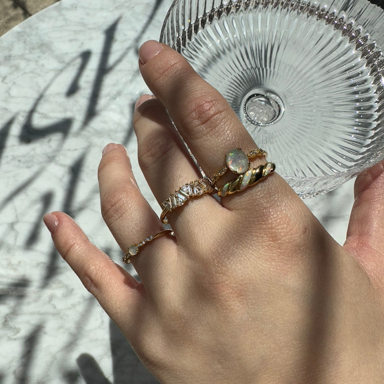 A hand adorned with four gold rings, including the Opal Sparkle Ring and twisted designs, holds a clear ribbed glass dish above a sunlit marble surface with soft shadows.