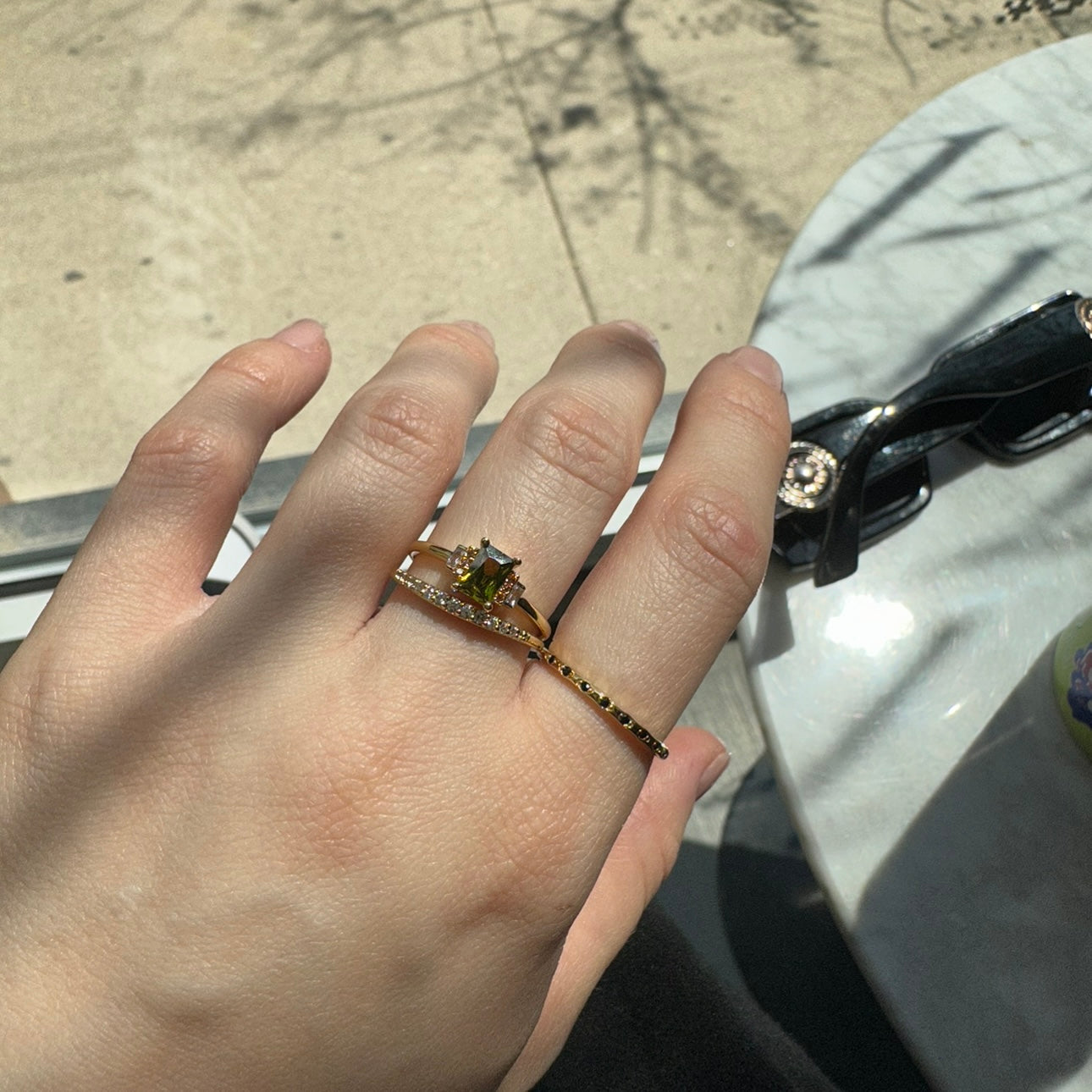 A hand with light skin tone wears the Regency Stacking Ring, featuring yellow gold plating and a green gemstone, along with a band ring. Both rest on white marble near black sunglasses, bathed in soft sunlight and shadows for a natural look.