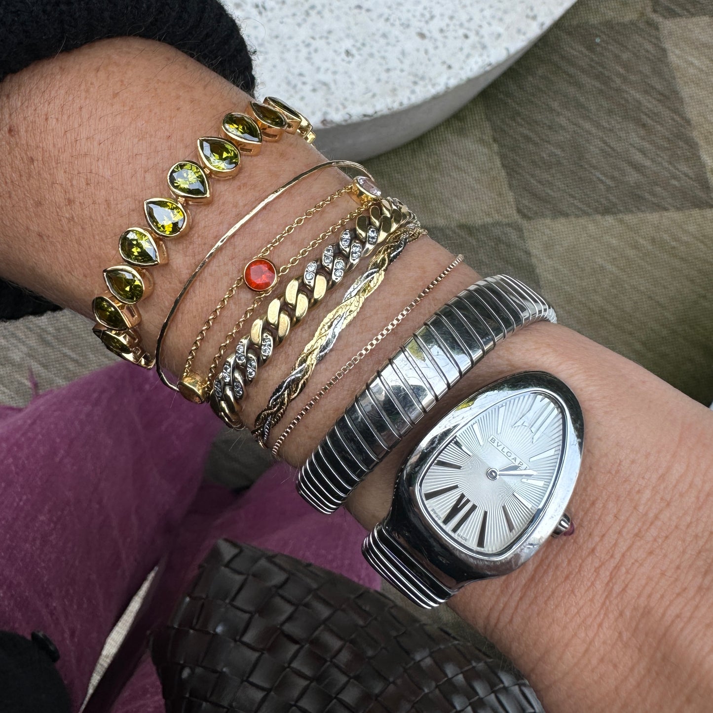 A wrist adorned with several gold bracelets, including the Solid Gold Mixed Metal Braided Bracelet, plus a silver watch with an oval face and Roman numerals. The person is wearing a dark sweater.
